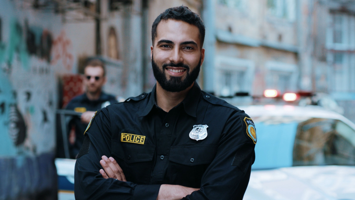 Smiling young man cops stand near patrol car look at camera enforcement happy officer police uniform auto safety security communication control policeman portrait close up slow motion.