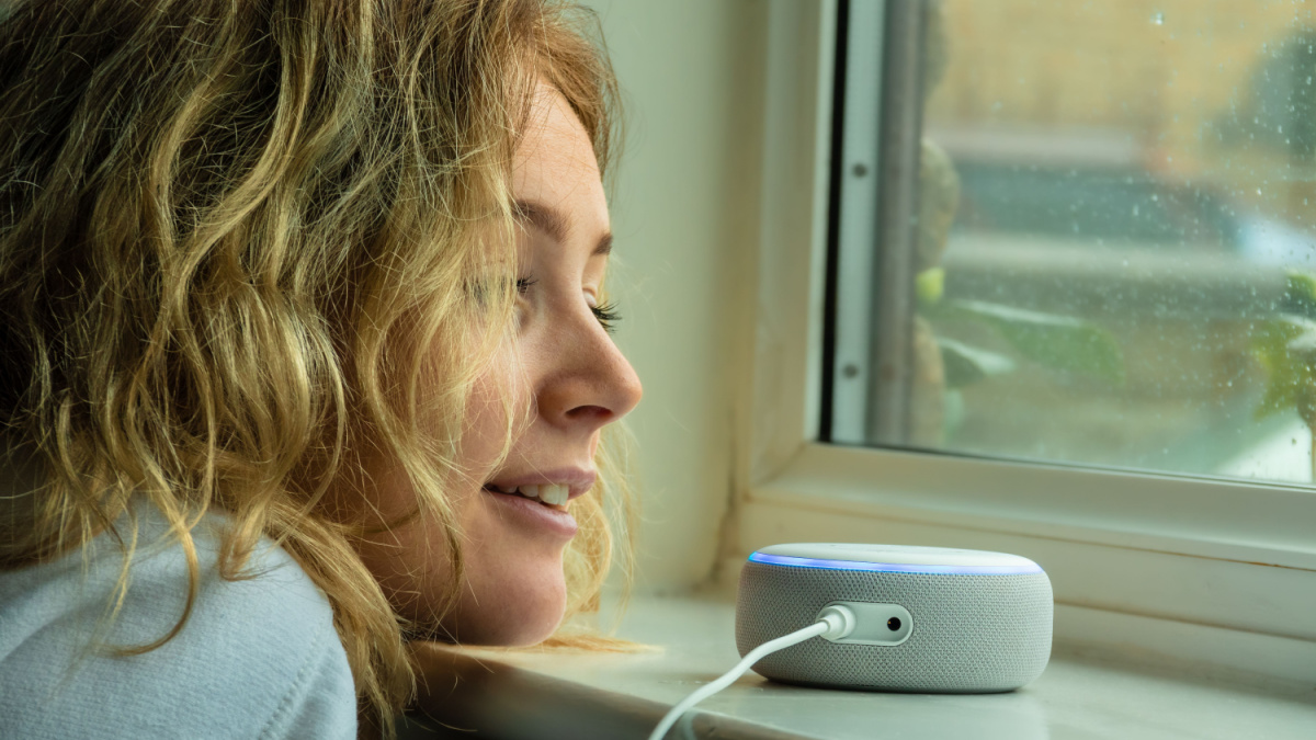 MOSTERTON ENGLAND FEBRUARY 2020 - Teenage girl talking to an Amazon Alexa Echo Dot by a window on a rainy day.