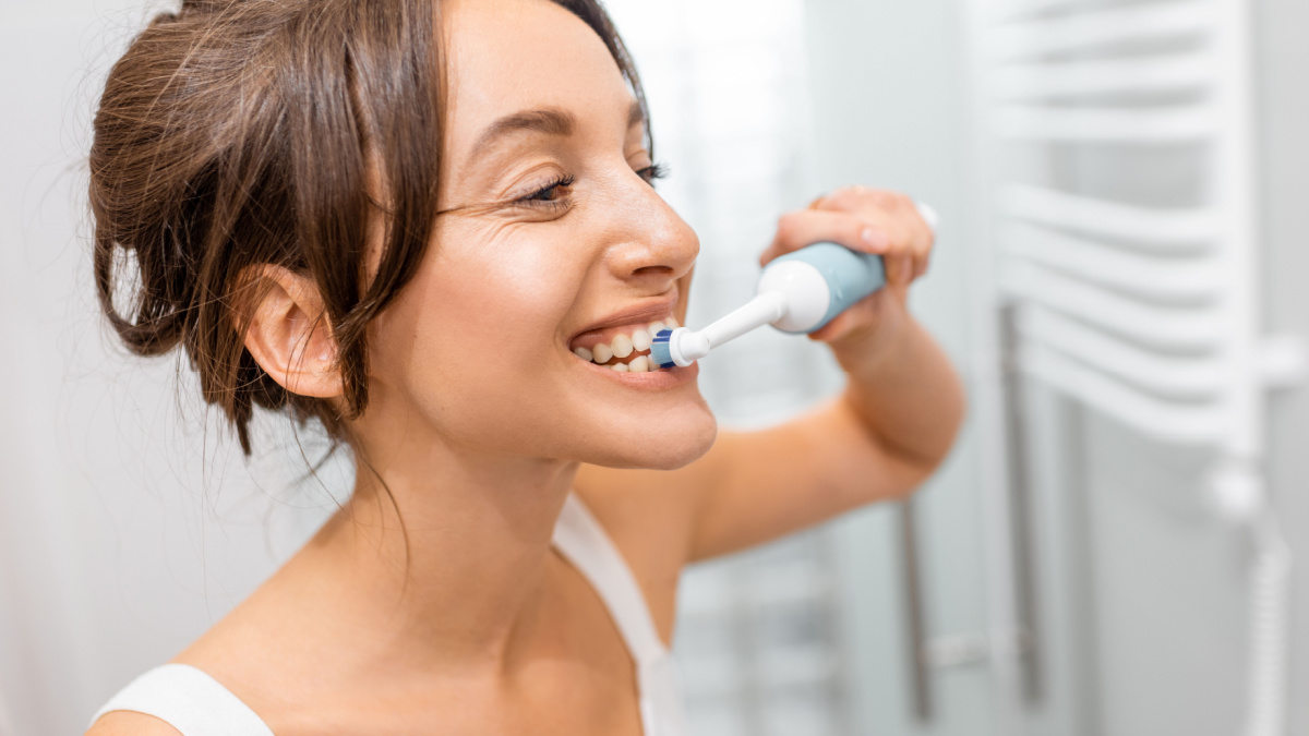 Young and cheerful woman brushing teeth with electric toothbrush during morning hygiene procedures in the bathroom