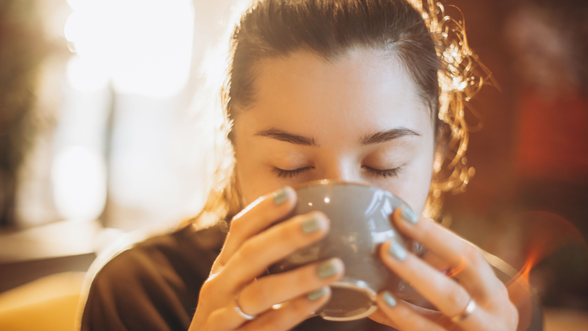 portrait of a beautiful woman drinking coffee or tea with a large mug sitting in a room where a lot of bright sunlight and sunlight go through the windows and create a warm light atmosphere.