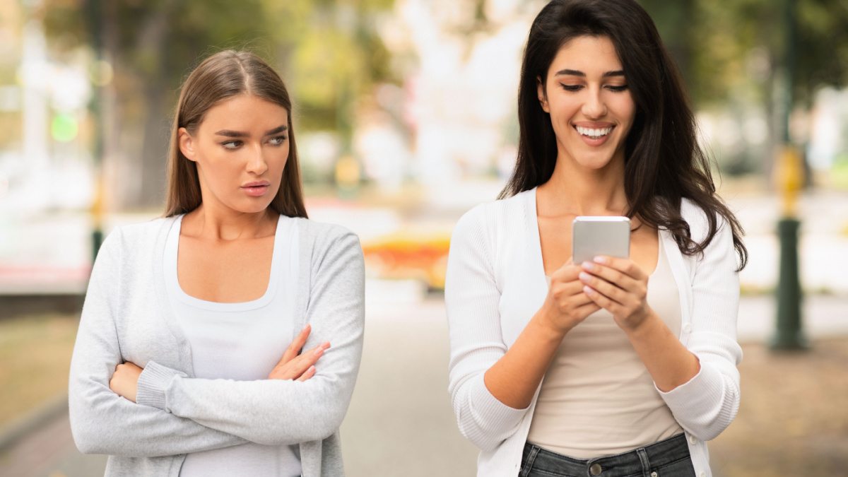 Envy Concept. Jealous Girl Looking At Friend's Phone Walking Together Outdoors At Park.
