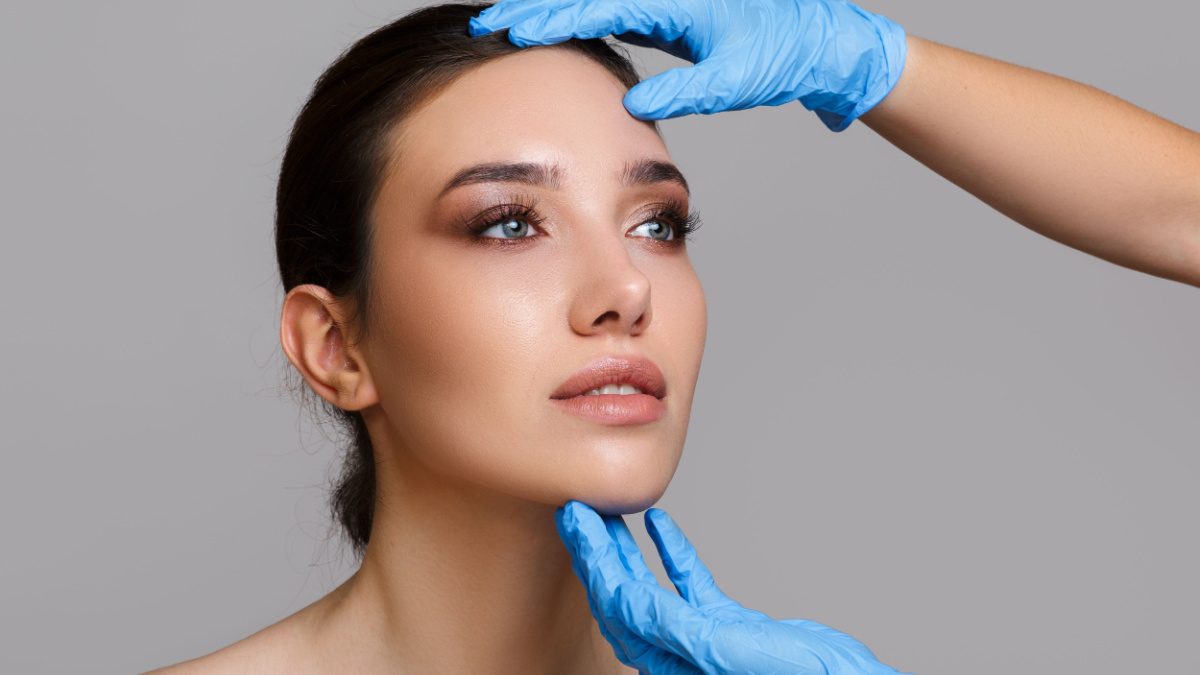 Facial beauty. Portrait of a beautiful young woman with perfect makeup and soft smooth skin. Close-up of beautician hands in gloves touching the face. Plastic surgery concept.