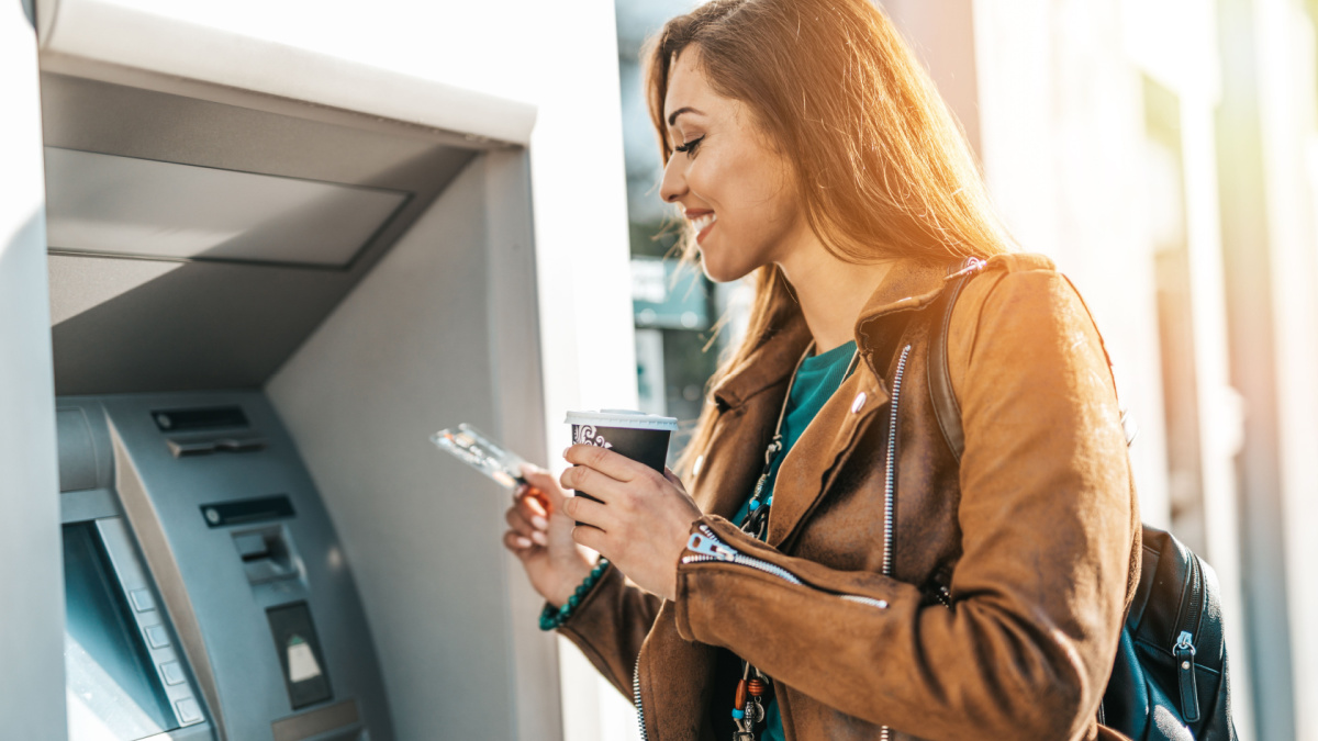 Happy young adult woman standing in front of ATM machine, smiling and holding credit or debit card.