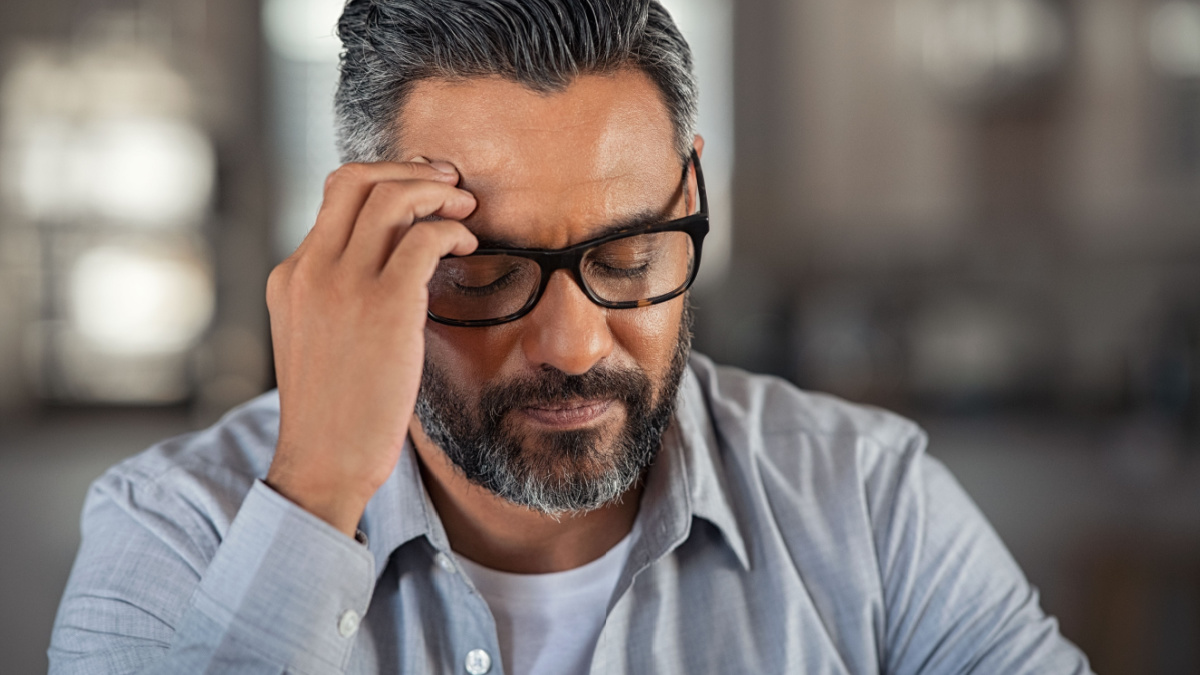 Frustrated middle aged man sitting on couch at home. Close up face of stressed indian businessman wearing eyeglasses with eyes closed.