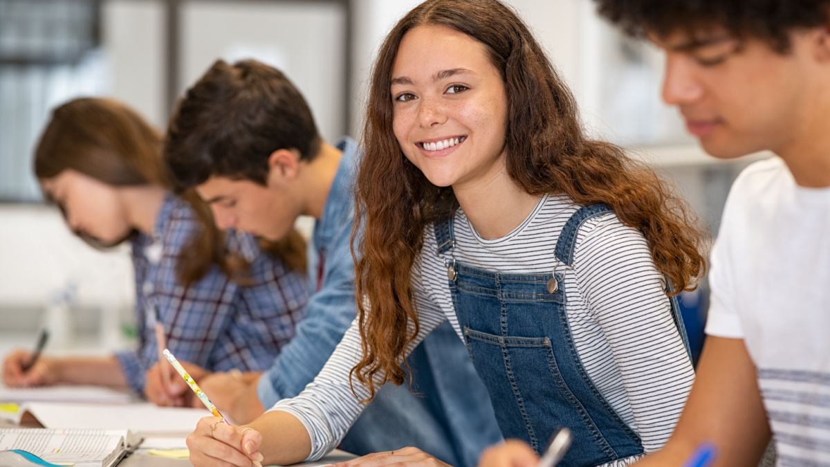 Satisfied young woman looking at camera. Team of multiethnic students preparing for university exam. Portrait of girl with freckles sitting in a row with her classmates during high school exam.