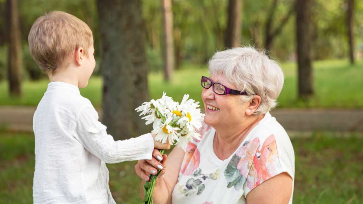 Beautiful boy giving a flower to grandma. Happy mothers day. Grandson and grandmother spending time together. Act of kindness to an elderly woman. Funny boy with flowers and his grandmother in park.