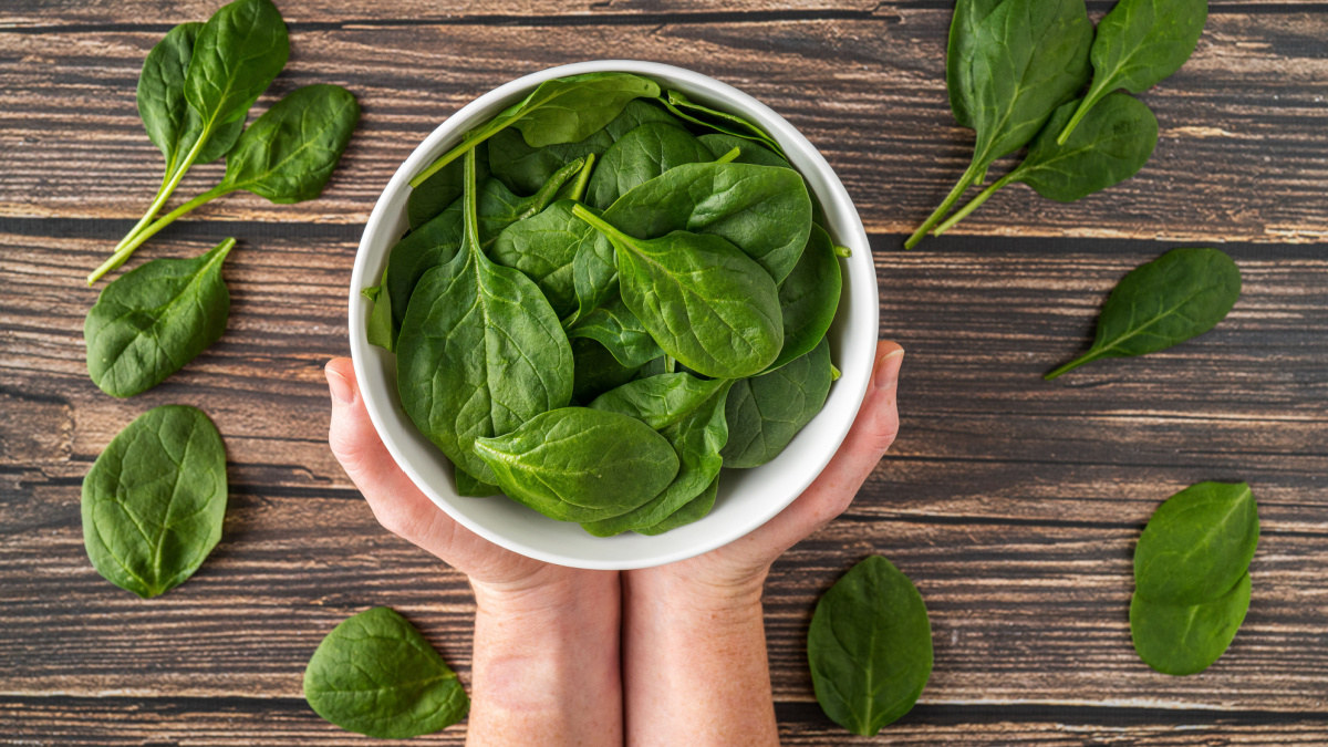 Fresh Organic Healthy Spinach in White Bowl Overhead Shot with White Female Hands Holding White Bowl on Wood Texture Table Background.