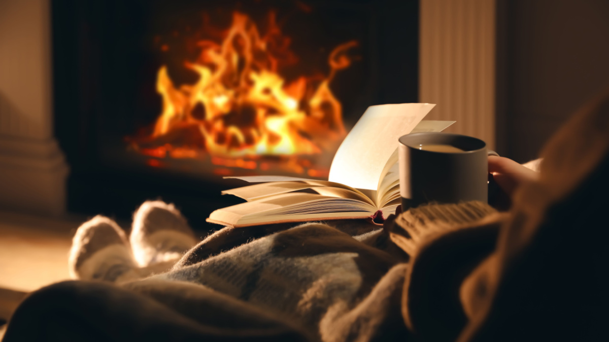 Woman with cup of drink and book near fireplace at home, closeup.
