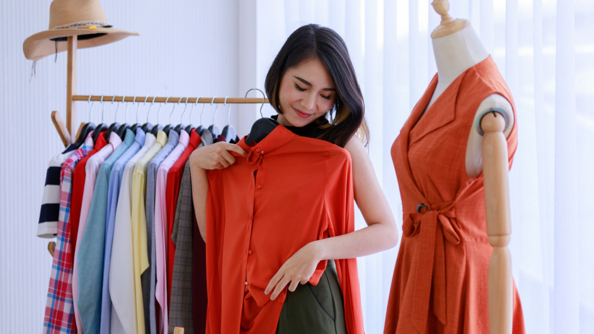 Smiling Asian woman Choose and try on clothe orange color in the Tailor shop. Fashion designer standing in clothing In order to repair, service for customers. Concept Profession Dressmaker designer