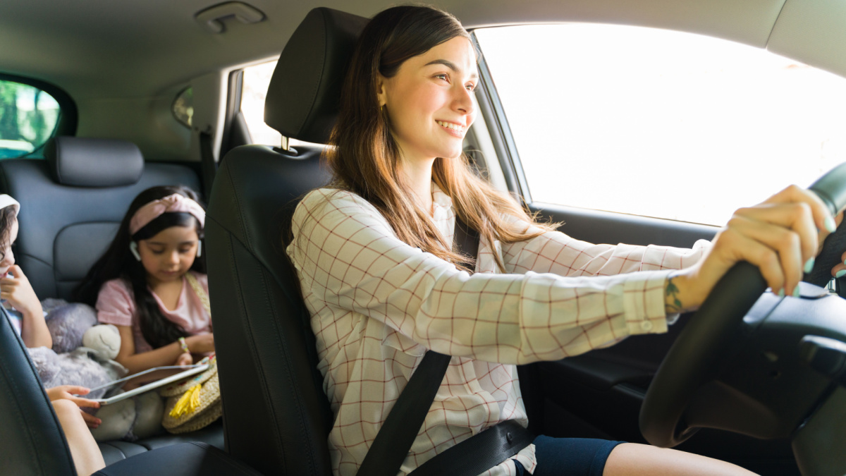 Entertaining my children. Beautiful young mom smiling while driving a car with her kids in the back watching a movie on a tablet with headphones during her errands.