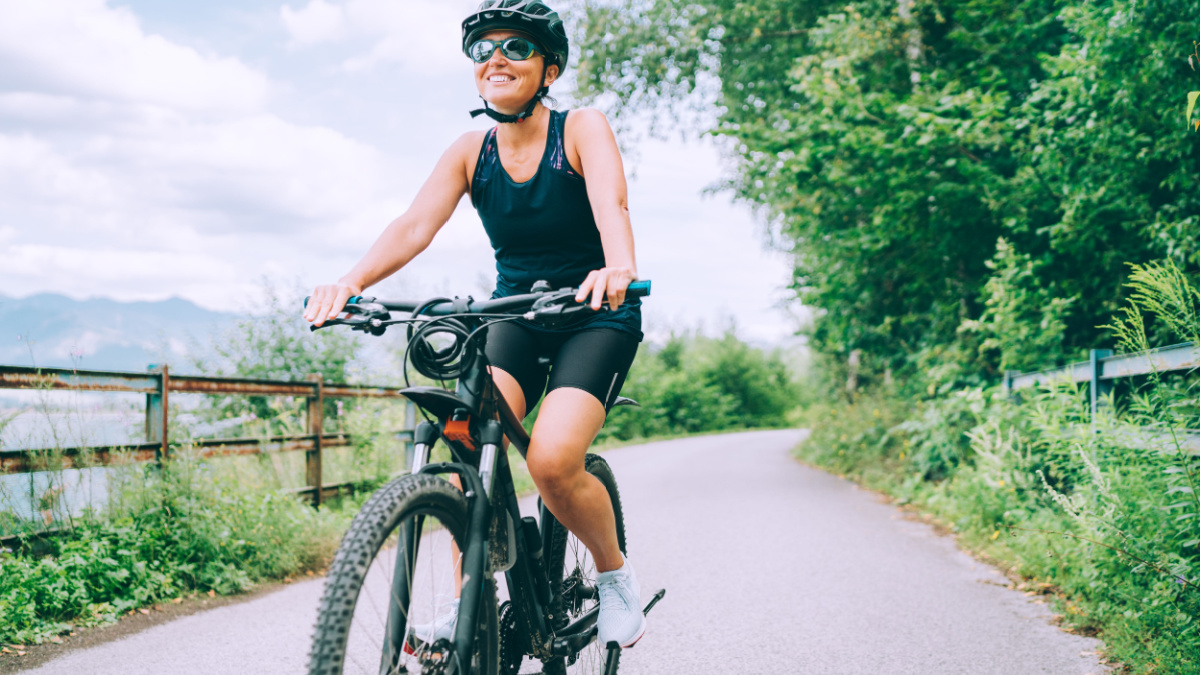 Portrait of a happy smiling woman dressed in cycling clothes, helmet and sunglasses riding a bicycle on the asphalt out-of-town bicycle path. Active sporty people concept image.