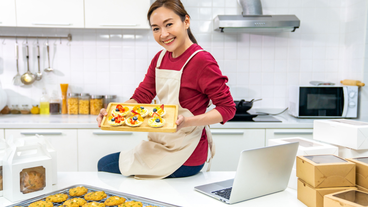 Portrait of Asian woman bakery shop owner using laptop computer advertising online bakery store on social media in the kitchen.