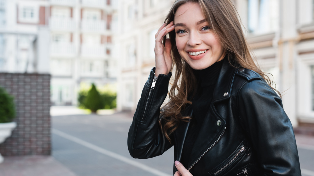 happy young woman in black turtleneck and leather jacket adjusting hair on urban street of europe