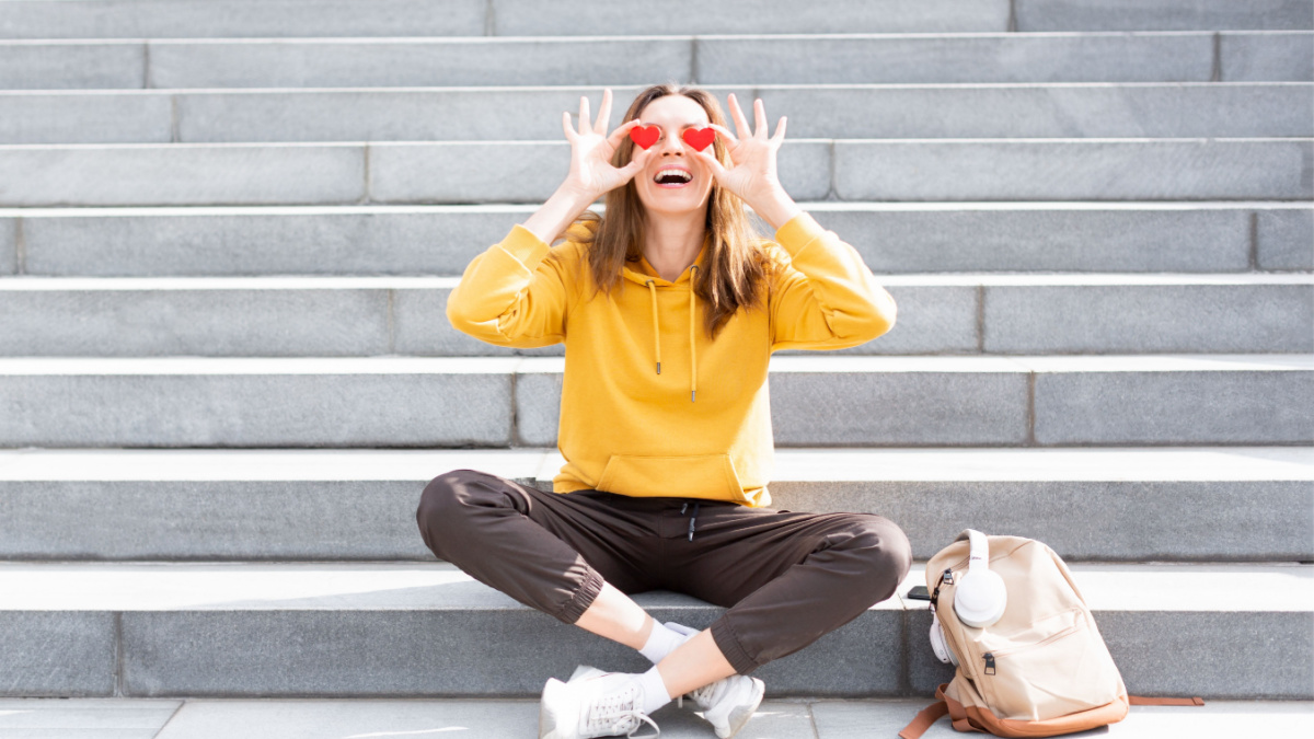 Cheerful, happy and optimistic person. Attractive expressive caucasian woman in casual wear sits on a stair steps with a backpack and looking at world using two small red hearts symbols