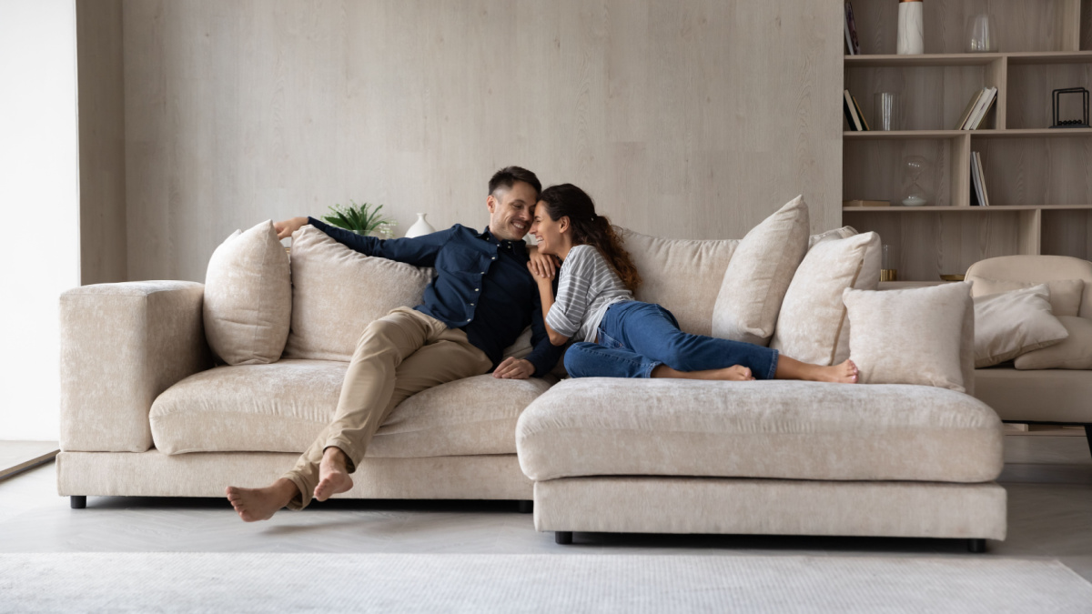 Full length happy relaxed young family couple homeowners or renters lying on big cozy sofa, enjoying carefree leisure weekend time, celebrating moving into new contemporary apartment, tenancy concept.
