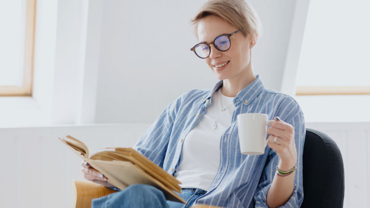 a young European woman with short blonde hair is drinking coffee or tea and reading a book. a woman is engaged in education or is resting.