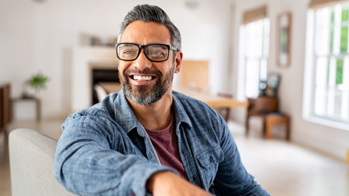 Happy mature middle eastern man wearing eyeglasses sitting on couch. Portrait of indian man relaxing at home and looking away with big smile. Mid adult guy with specs thinking about his future.