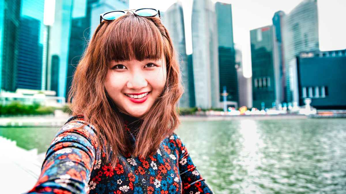 Young asian woman taking happy selfie traveling at Singapore skyline - Wanderlust life style concept with millenial girl having fun by urban city surrounds - Vivid bright filter with focus on face.