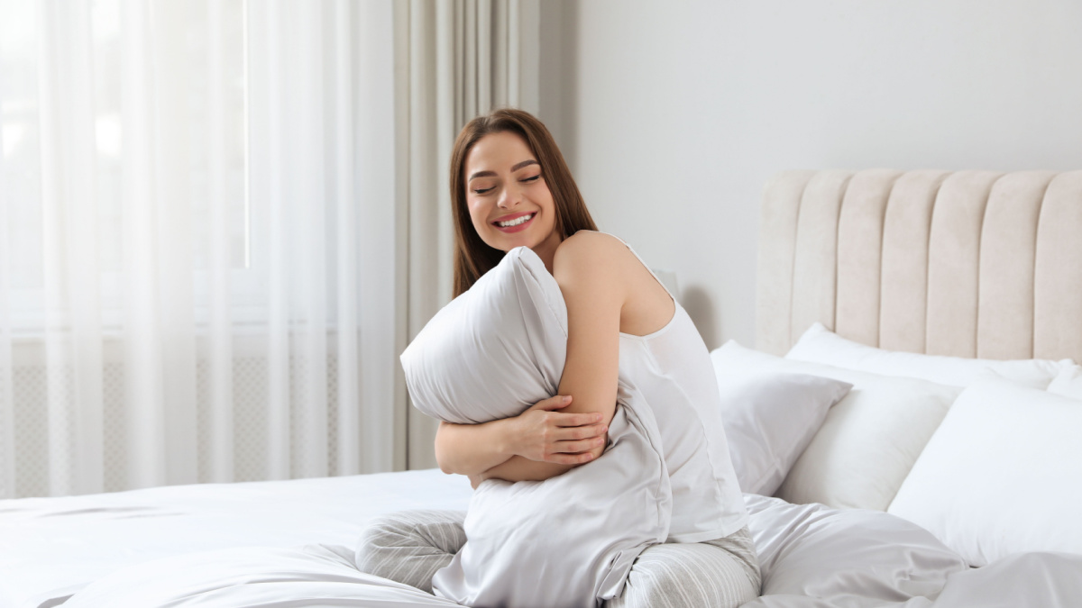 Young woman hugging pillow on comfortable bed with silky linens