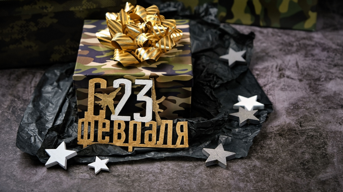 gift box with military camouflage pattern and golden bow, happy February 23 russian text on dark background.
