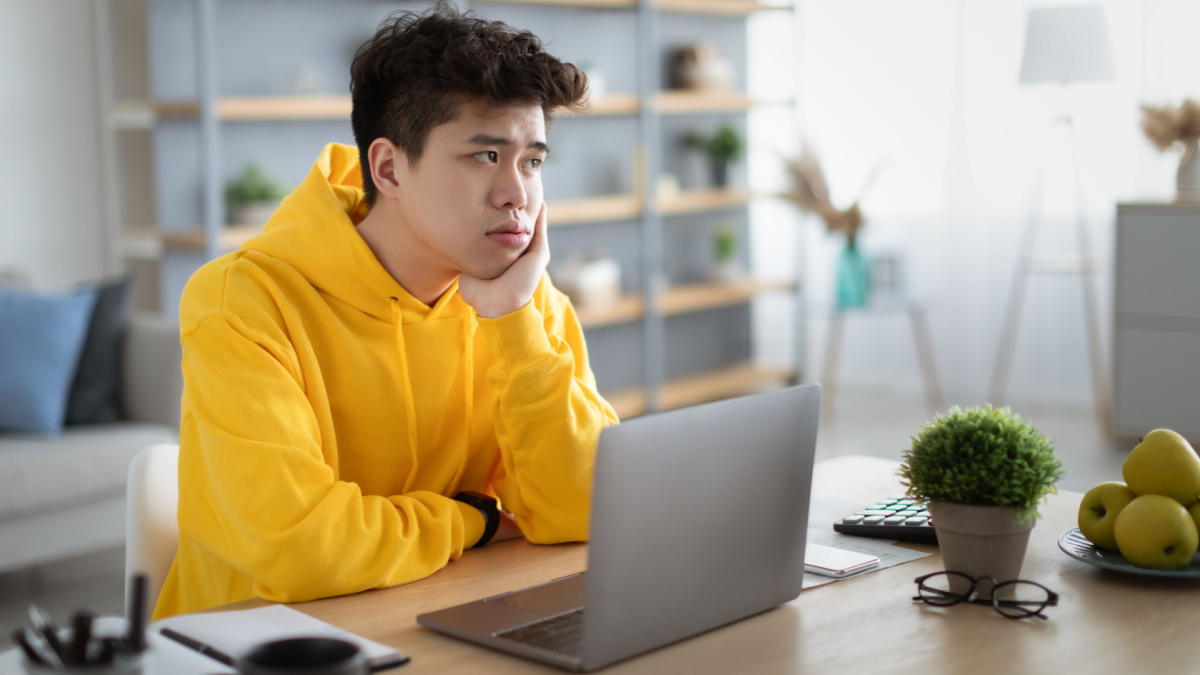 Bored young Asian guy sitting at desk using pc laptop, looking aside, thinking about something during online lesson or remote work at home office. Tired man having dull distant job, feeling lazy