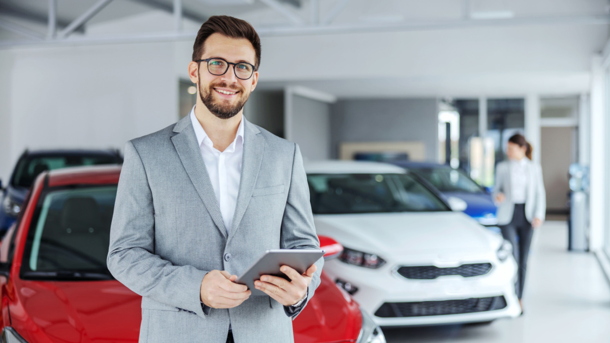 Smiling friendly car seller in suit standing in car salon and holding tablet. It's always pleasure to buy a car on a right place.