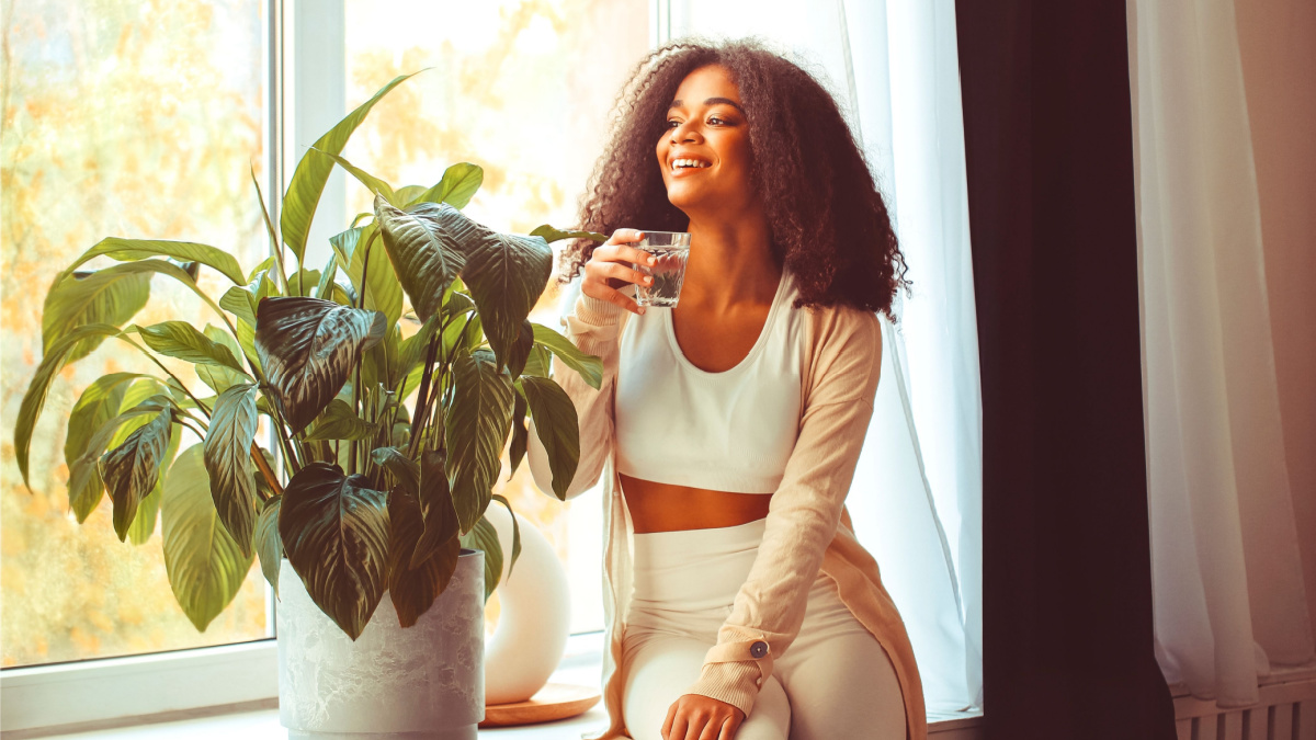 Young positive african american woman sitting on windowsill with glass of pure mineral water and looking through window with smile, drinking aqua in morning, starting new day with healthy habits.