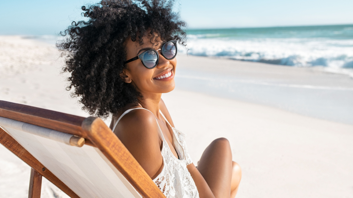 Portrait of happy young black woman relaxing on wooden deck chair at tropical beach while looking at camera wearing spectacles.
