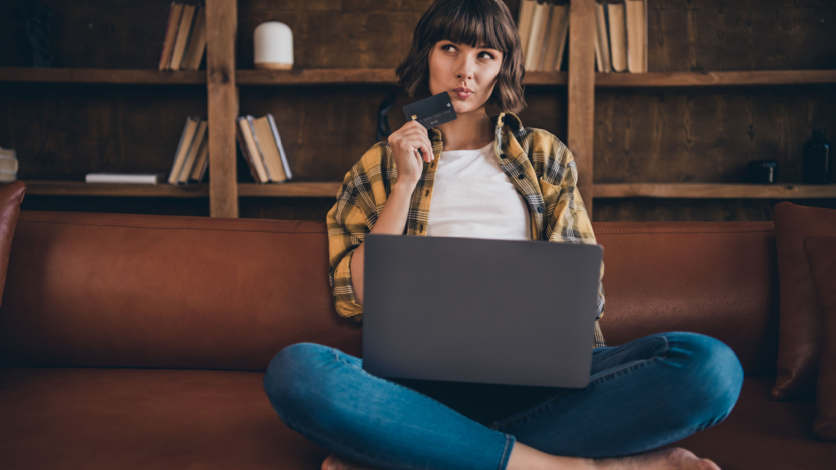 Full size photo of thinking young bob hairdo lady sit with laptop hold card wear shirt jeans bare foot indoors alone.
