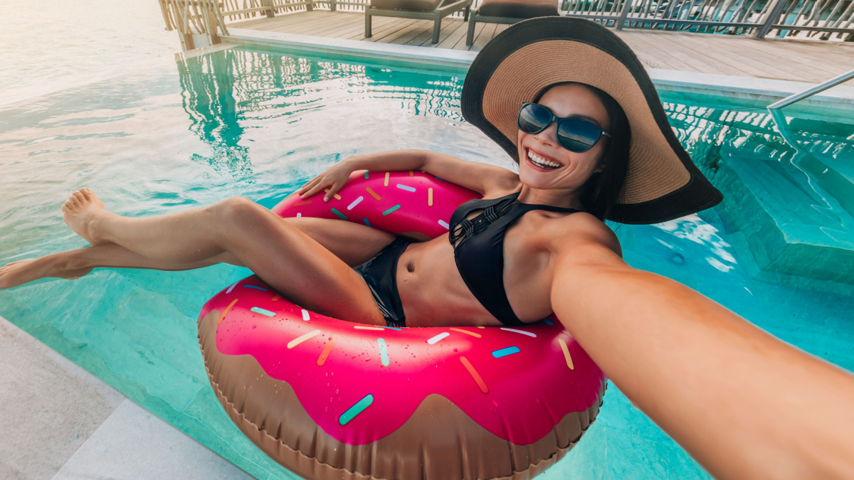 Relaxing woman floating in donut inflatable swimming pool toy at luxury resort taking selfie photo using sunbathing. Travel vacation hotel lifestyle.