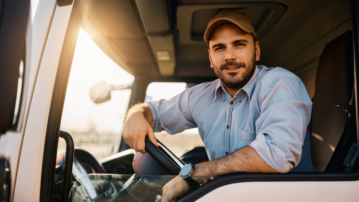 Young truck driver sitting behind steering wheel in a cabin and looking at camera.
