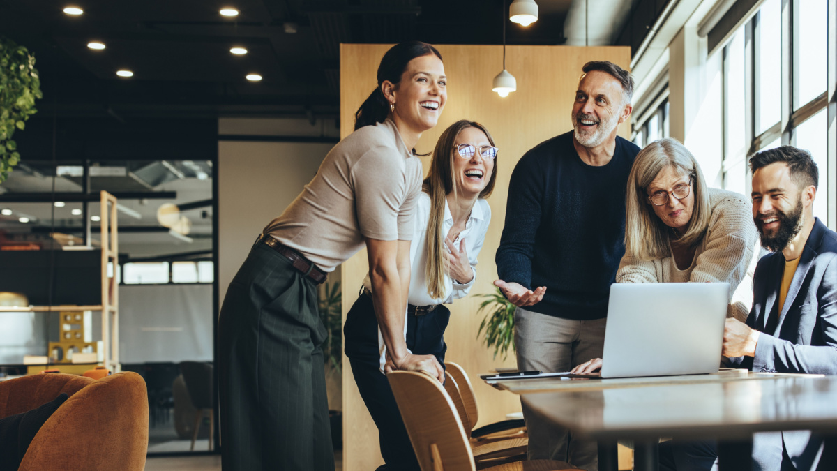Happy businesspeople laughing while collaborating on a new project in an office. Group of diverse businesspeople using a laptop while working together in a modern workspace.