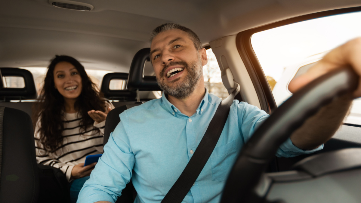 Nice Ride. Portrait of happy male driver riding car looking at cheerful beautiful lady sitting inside auto on back passenger seat, female using cell phone and talking with guy, windshield view.