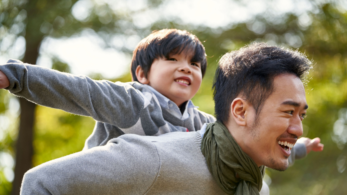 young asian father carrying son on back having fun enjoying nature outdoors in park.