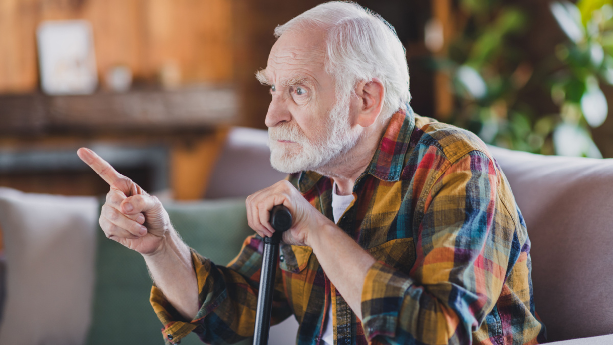 Photo of nervous angry retired man dressed plaid shirt holding nursery cane pointing finger sitting couch indoors apartment