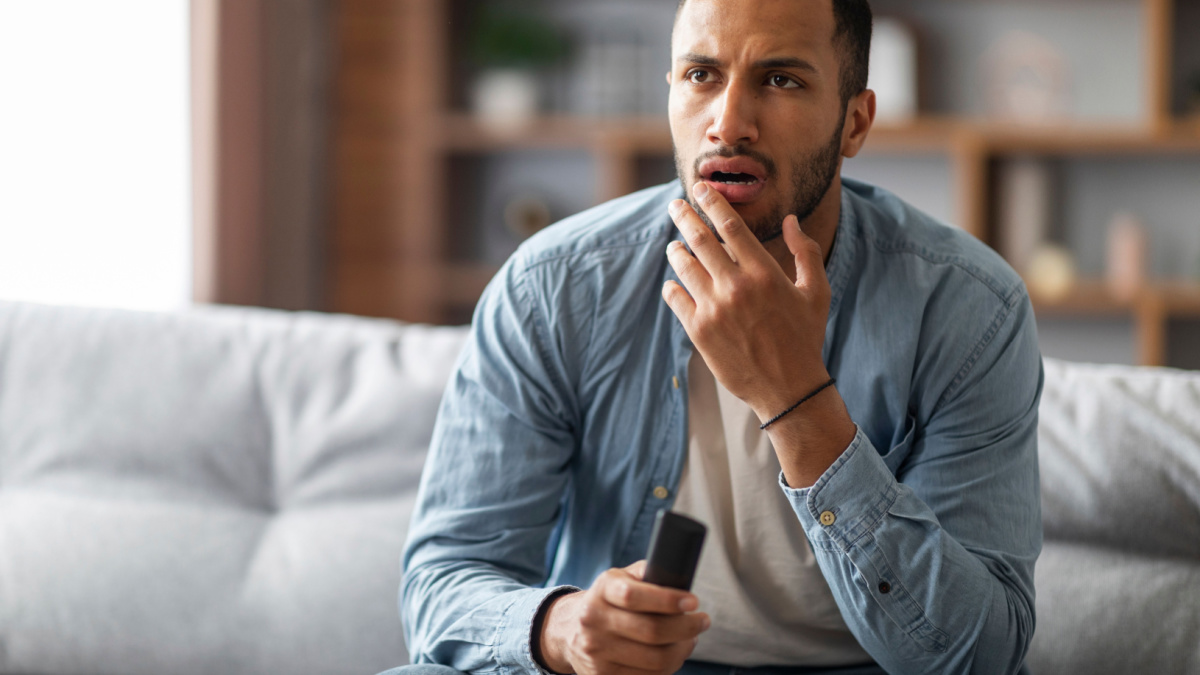 Shocked Black Man With Remote Controller In Hand Sitting On Couch At Home, Confused Young African American Guy Watching Tv Show In Living Room, Emotionally Reacting To Shocking Content, Copy Space