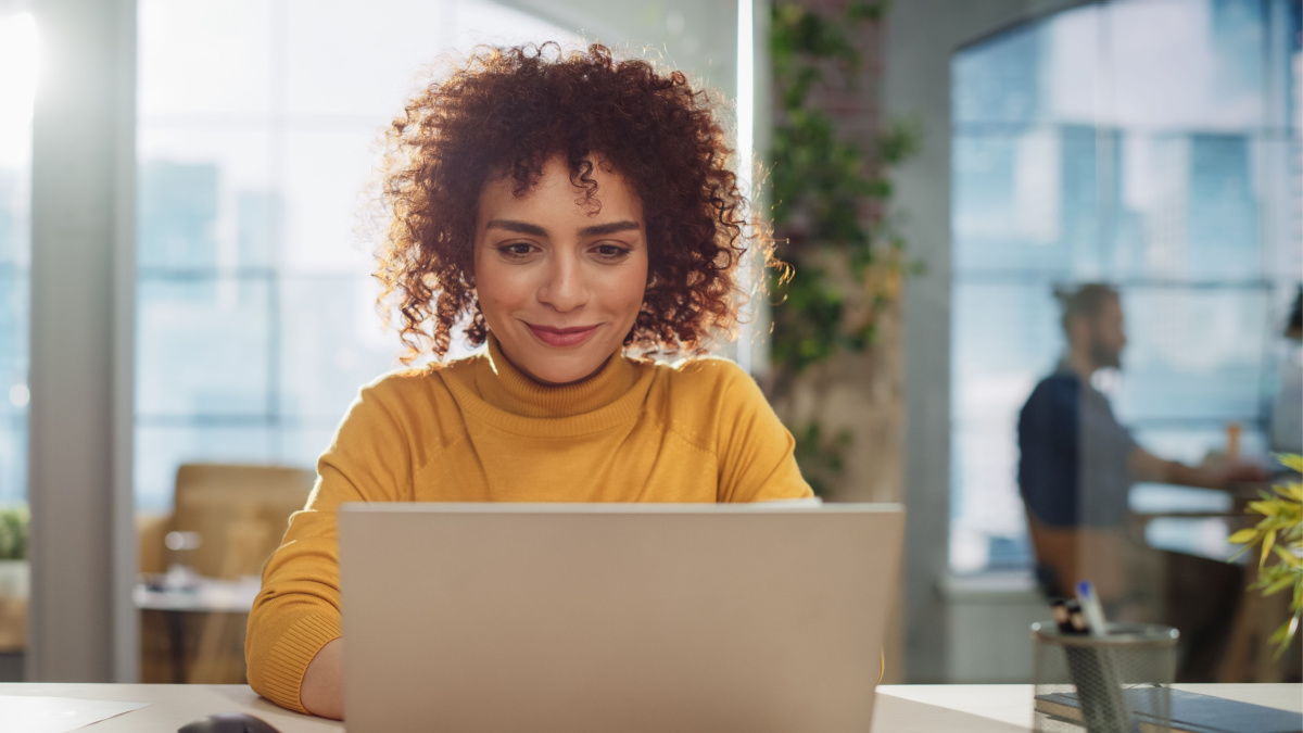 Beautiful Middle Eastern Manager Sitting at a Desk in Creative Office. Young Stylish Female with Curly Hair Using Laptop Computer in Marketing Agency. Colleagues Working in the Background.