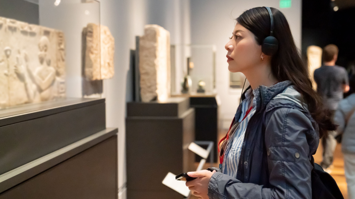 side view of asian Korean college girl taking audio tour in the history museum located in los angeles usa. she is looking at an ancient carving displayed in glass case