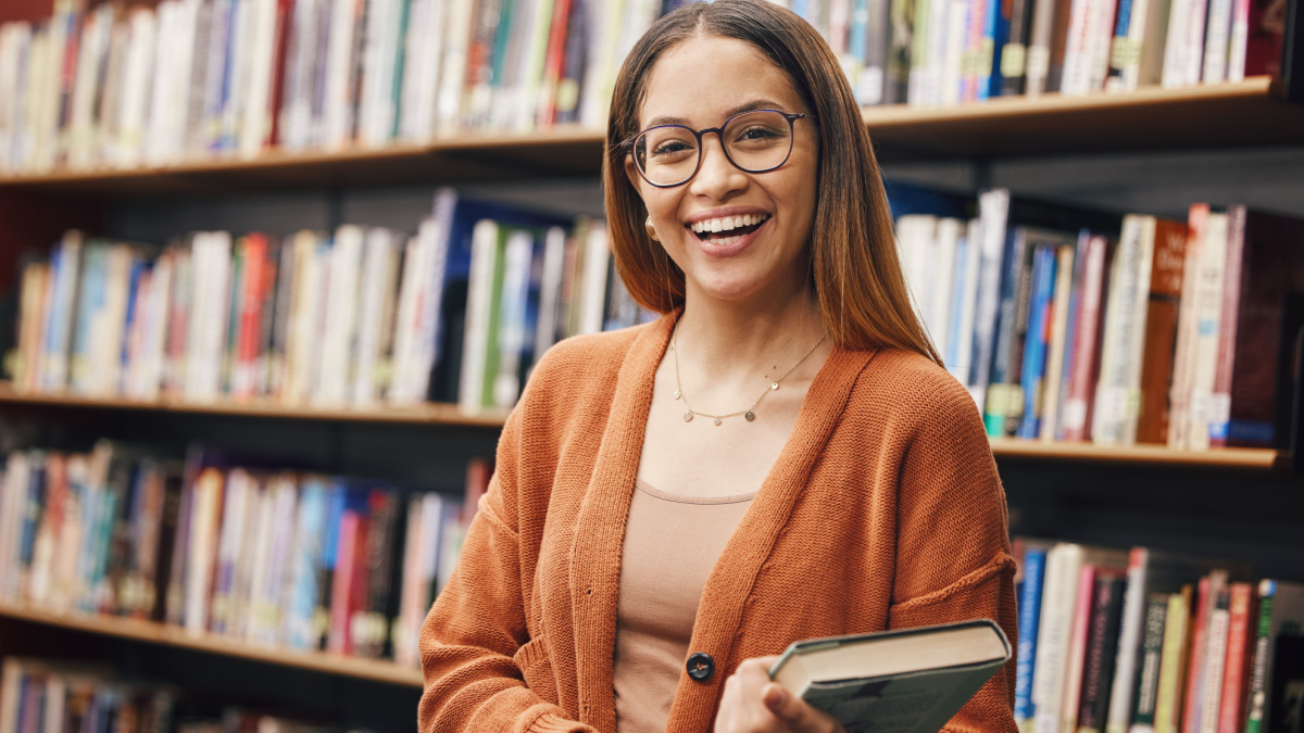 Face, student and woman in university in library ready for learning. Portrait, education and happy female from Brazil standing by bookshelf with book for studying, knowledge and literature research.