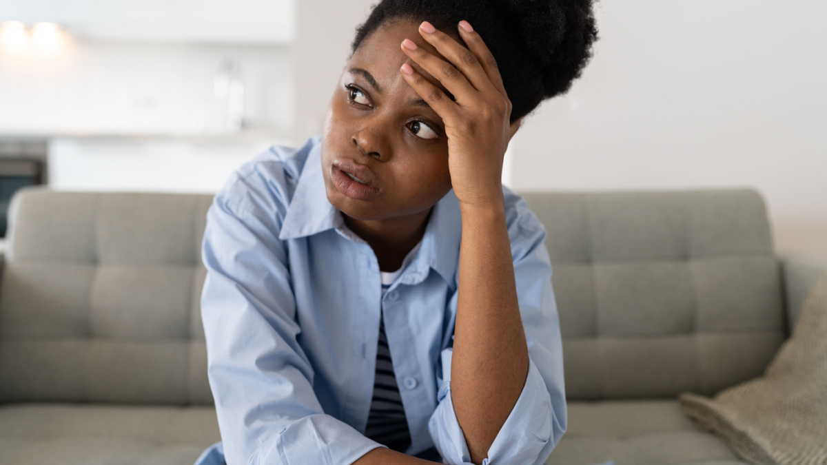Flustered African American woman very nervous sighing and touching face with hands sits on couch of home.