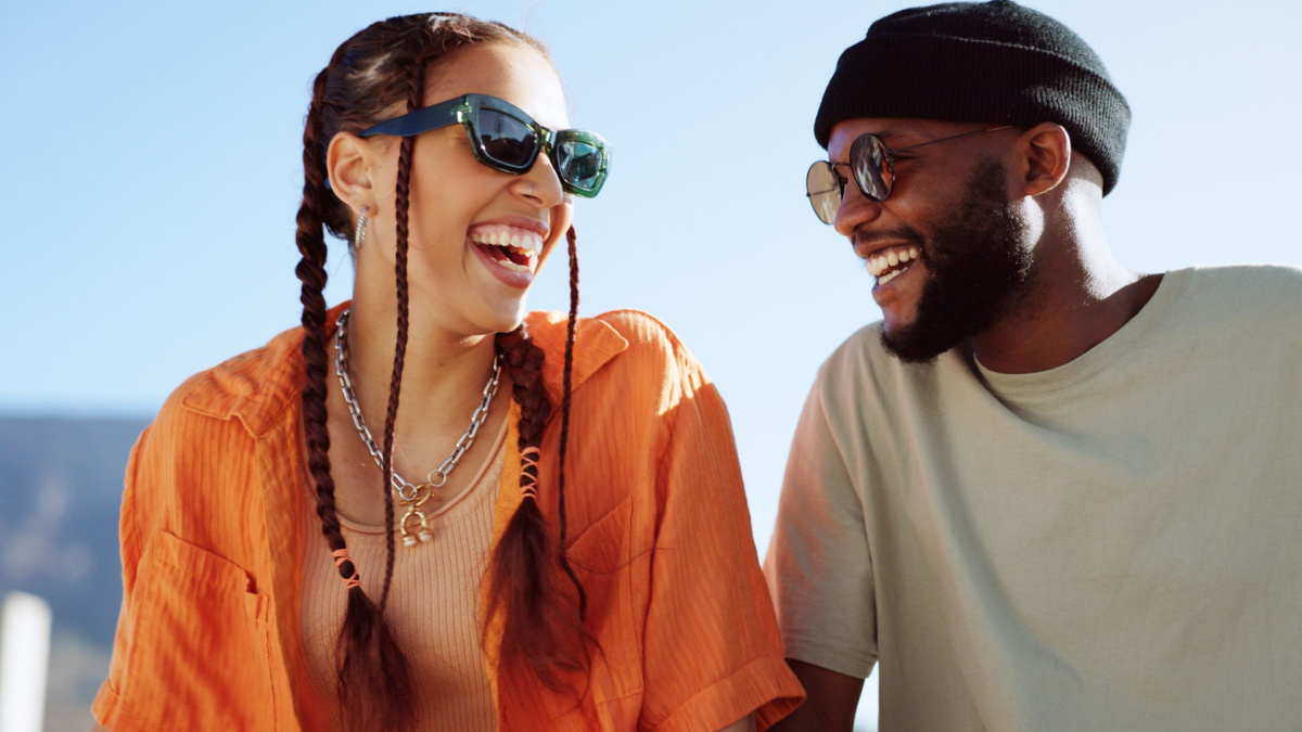 Friendship, flirting and couple with sunglasses in city sitting on building rooftop laughing. Love, friends and romance, urban dating and freedom for gen z woman and man with smile on crazy fun date.