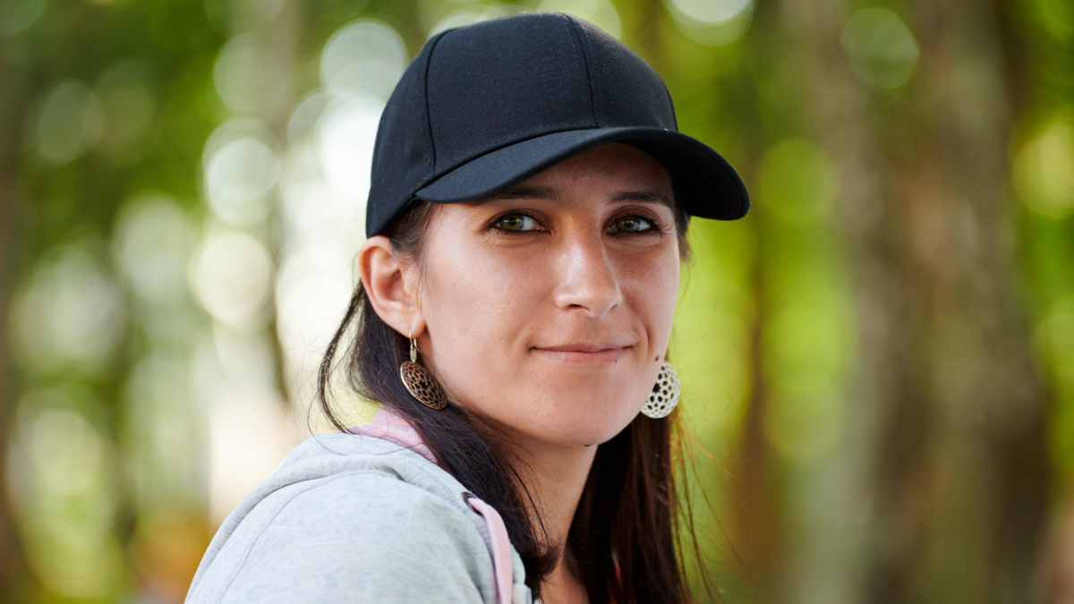 Young woman with embarrassed smile in black cap and gray hoodie on outdoor green forest bokeh background.