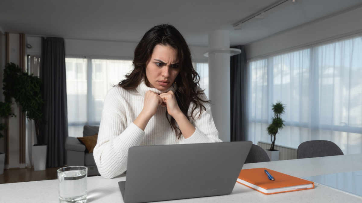 Young shocked businesswoman annoyed unpleasant spam message pop-up ad frantically looking at laptop screen while sitting and working from home trying to cover monitor with hands, inappropriate content