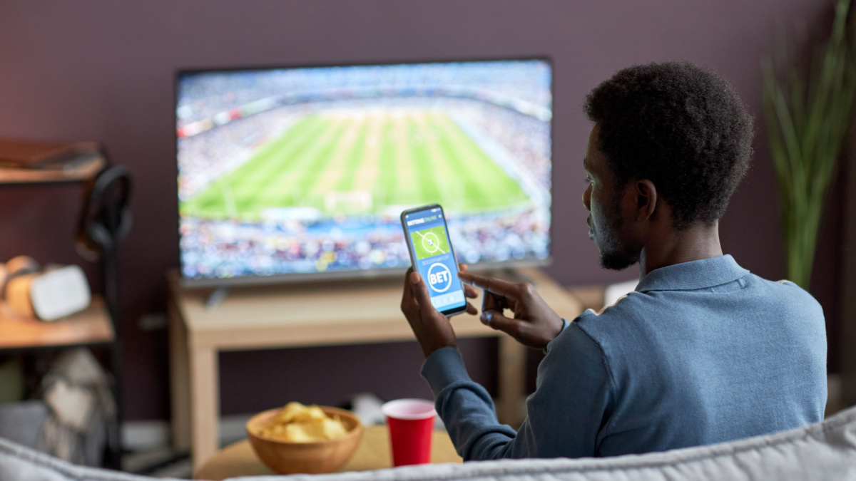 Back view of man holding smartphone with sports bets app on screen while watching football match at home.