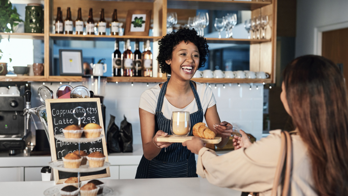 Happy woman, barista and serving customer at cafe for service, payment or order on counter at coffee shop. African person, waitress or employee in small business restaurant helping client at checkout