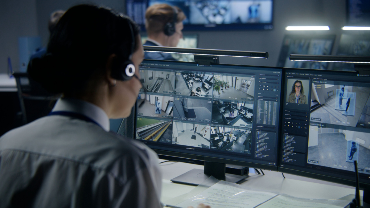 Diverse employees work in security control center, watch CCTV cameras with AI face scanning. Female operator uses PC. Computer monitors and big digital screens with surveillance cameras video footage.
