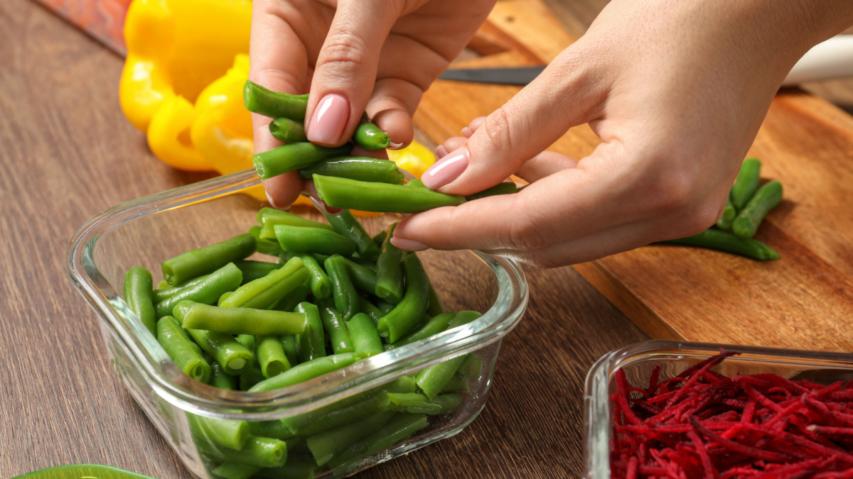 Woman putting green beans into glass container at wooden table, closeup. Food storage