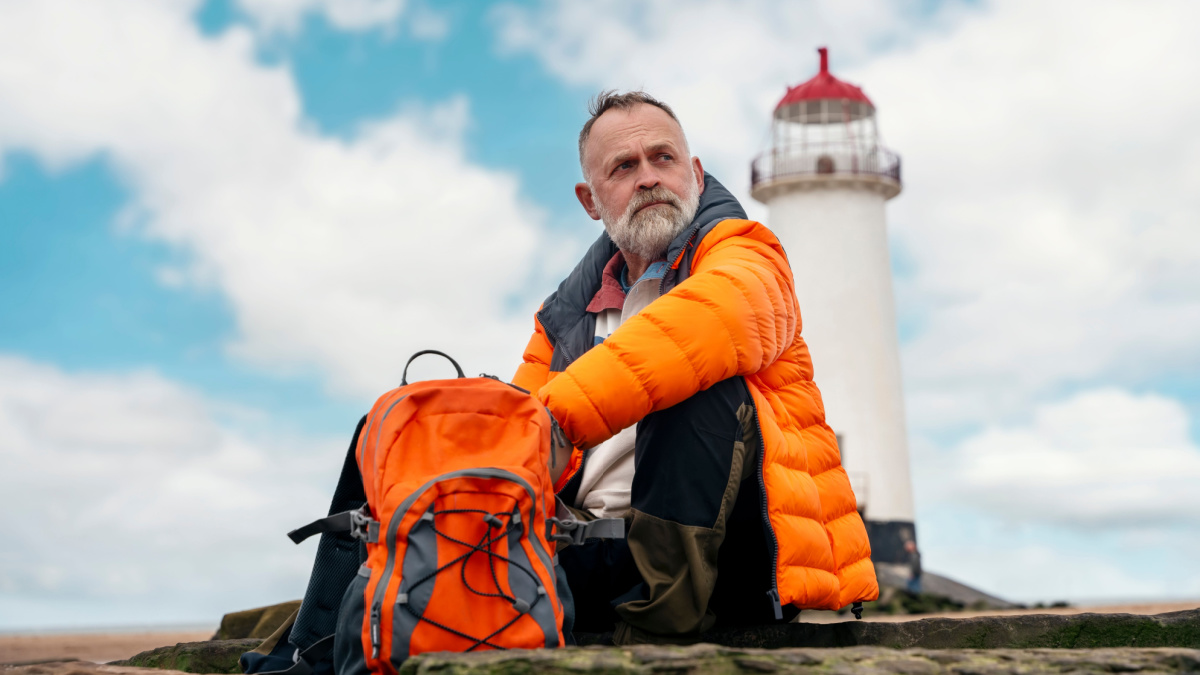 old man enjoying life at full by travelling around world sittig on the seaside against lighthouse on sunny Autumn day.