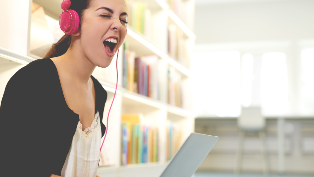 Funny happy female student sitting on the floor and singing while listening to loud music through pink headphones connected to laptop, in the interior of a public academic library