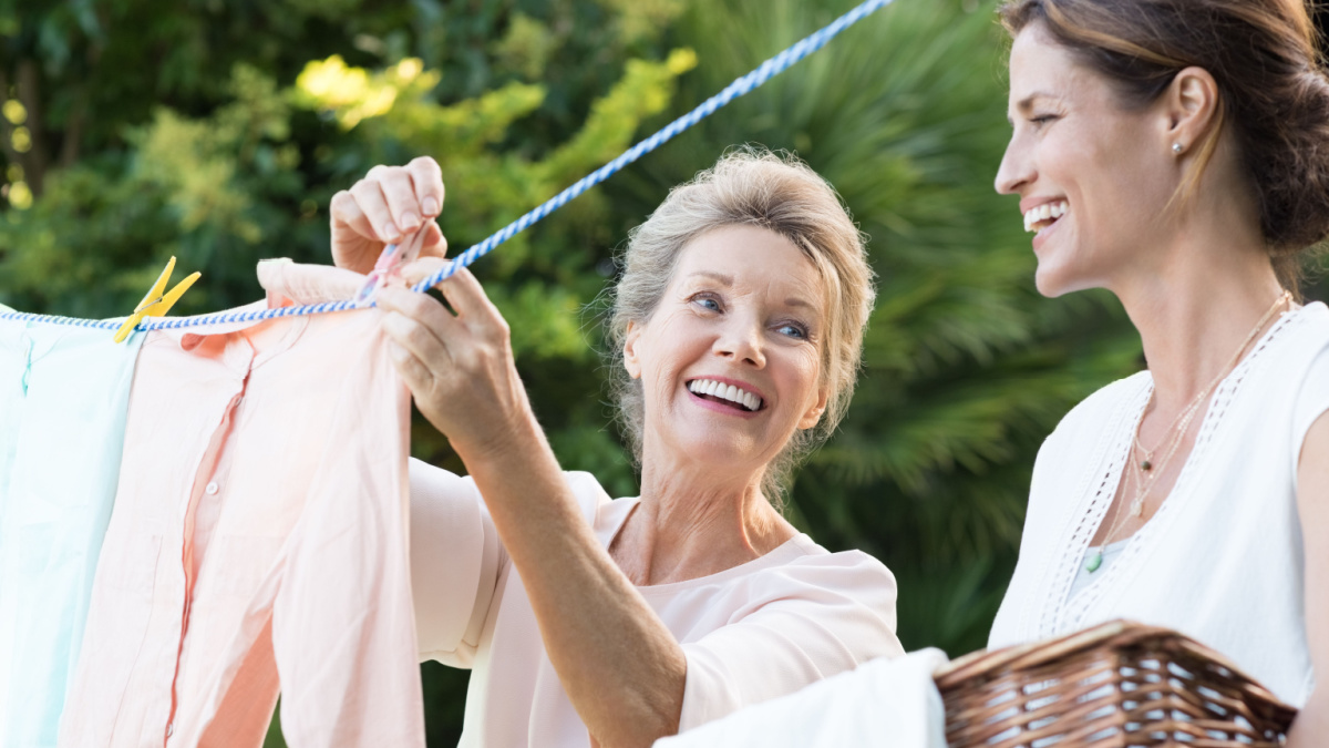 Older mother and young daughter hanging clothes outdoor to dry. Smiling daughter helping mother with laundry. Cheerful mother and daughter in conversation while hanging clothes outside.